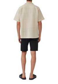 Korte mouwen, crèmekleurig shirt met een gestructureerd open breiontwerp, gecombineerd met zwarte shorts en instappers, gezien van achteren.