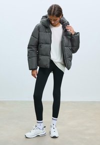 Dark gray puffer jacket with a hood, quilted design, worn over a white shirt and black leggings. Model stands in white sneakers with stripes.