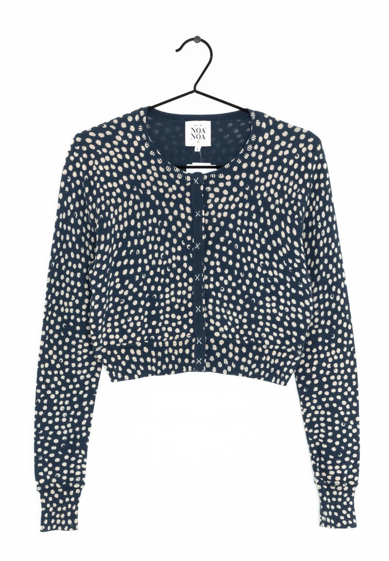 Cardigan court bleu marine avec motif de points blancs irréguliers, manches longues, col rond et fermeture par boutons, accroché sur un cintre noir.