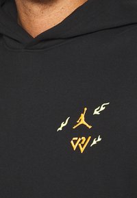 Sudadera negra con bolsillo tipo canguro. Presenta un logo Jumpman en naranja y detalles en llamas blancas, fabricada con un suave material de mezcla de algodón.