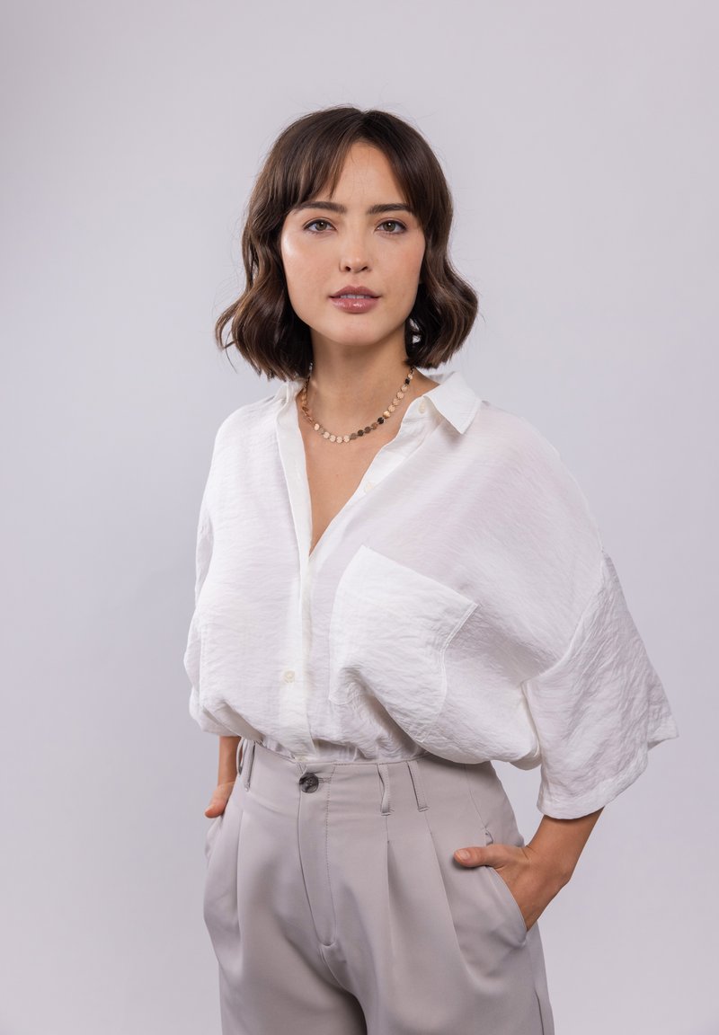 Weißes, strukturiertes Button-Down-Hemd mit einer einzelnen Tasche, kombiniert mit hellgrauen, gefalteten Hosen. Schlichter Ausschnitt, lässige Passform.