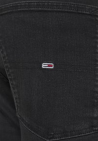 Los pantalones de mezclilla negra muestran un primer plano del bolsillo trasero con un pequeño logotipo bordado en blanco, rojo y azul. La tela tiene una superficie ligeramente texturizada.