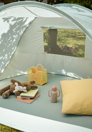 Raidallinen leikkiteltta, jonka sisällä on pehmeä keltainen tyyny, vaaleanpunavalkoinen vesipullo, keltainen kori ja pehmoleluja vihreällä matolla.