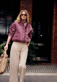 Gesteppte, violette Jacke mit hohem Kragen, kombiniert mit cremefarbenen weit geschnittenen Hosen. Hält eine strukturierte beige Tasche. Urbaner Hintergrund.