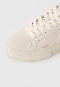 Sneaker in suede beige chiaro con lacci crema, dotato di una suola in gomma testurizzata e di un accento dorato del logo sul lato.