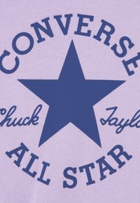 Un tissu violet clair présente un logo étoile bleu marine foncé et un texte indiquant "CONVERSE ALL STAR" et "Chuck Taylor" en caractères gras et cursifs.
