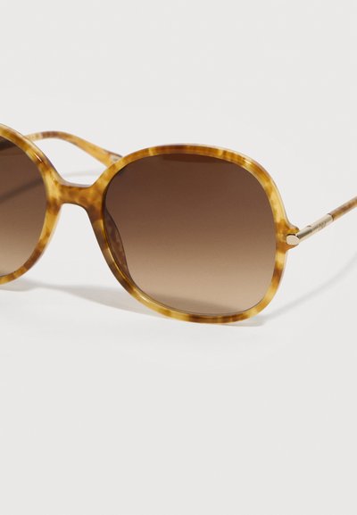 Στρογγυλά γυαλιά ηλίου με καφέ διαβάθμιση και λεπτά μπροστινά πλαίσια από ασετάτ με σχέδιο χελώνας, καθώς και μεταλλικούς βραχίονες, σε λευκό φόντο.