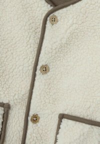 Veste en polaire beige clair avec une surface texturée, des bordures marron et des boutons ronds avec un design floral. Comprend deux poches avant.