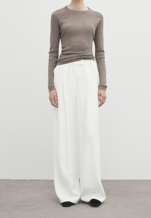 Personne portant un haut marron côtelé à manches longues ajusté et un pantalon blanc à jambes larges, debout sur un sol gris devant un mur blanc.