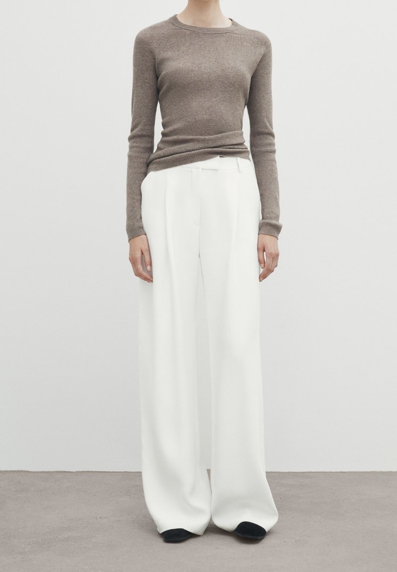 Personne portant un haut marron côtelé à manches longues ajusté et un pantalon blanc à jambes larges, debout sur un sol gris devant un mur blanc.