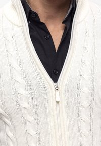 Lichtbeige kabelgebreide cardigan met een hoge kraag, ritssluiting en geribbelde structuur, gedragen over een zwart overhemd met knopen.