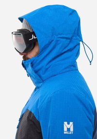 Blaue und schwarze wasserdichte Jacke mit strukturiertem Oberflächen, verstellbarer Kapuze und Logo am Ärmel, getragen mit Skibrille.