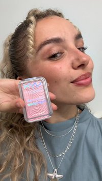 Un compact rettangolare trasparente con prodotto rosa e texture ondulata. La custodia trasparente presenta un coperchio blu con testo olografico.