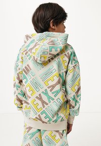 Mikina s kapucí vyrobená z měkké látky, která má vícebarevný geometrický vzor v tyrkysové, žluté a hnědé barvě. Rybí manžety a lem.