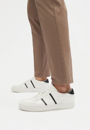 Beli usnjeni športni čevlji z črnimi črtastimi poudarki, ravnimi podplati in črno oznako na zadnjem delu. Ujemajo se z rjavimi hlačami, osredotočeni na podrobnosti obutve.