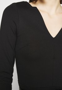 Haut noir à manches longues avec un col en V, texture de tissu lisse et coutures discrètes le long de la poitrine et des côtés.