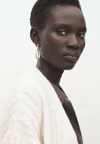 Primer plano de una persona que lleva un cárdigan blanco de punto con trenzas y una camisa marrón. Entre las características destacadas están un pendiente plateado en forma de gota y la piel con textura.