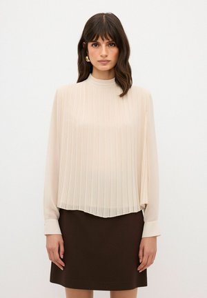 Beige geplooide blouse met een hoge hals en lange mouwen, gecombineerd met een bruine rok. De stof is licht en vloeiend met een zachte textuur.