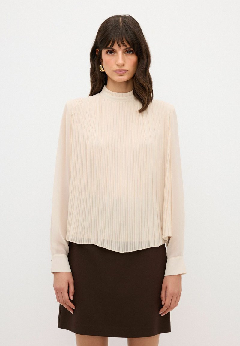 Blusa beige plissettata con colletto alto e maniche lunghe, abbinata a una gonna marrone. Il tessuto è leggero e fluido con una texture liscia.