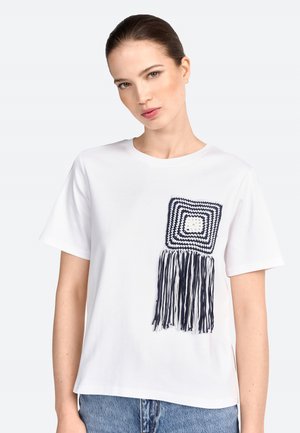 T-shirt en coton blanc avec une poche carrée en crochet bleu et blanc, équipée de longues franges sur le côté gauche. Coupe décontractée, manches courtes.