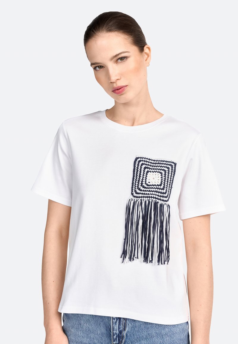 Hvid bomulds t-shirt med en blå og hvid hæklet firkantlomme med lange frynser på venstre side. Løs pasform, korte ærmer.