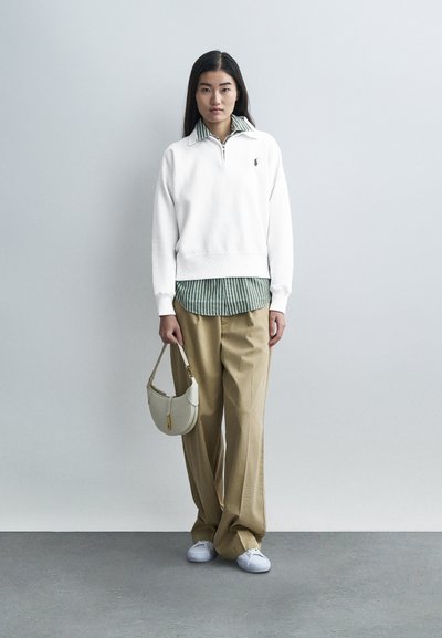 Sudadera blanca con cuello con cremallera, sobre una camisa a rayas verdes y blancas. Pantalones beige holgados y un bolso pequeño de color claro. Zapatillas blancas.