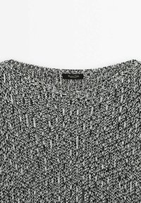 Schwarzer und weißer gestrickter Pullover mit weitem Halsausschnitt, der ein strukturiertes Muster und ein Etikett am Kragen aufweist, aus weichem Material gefertigt.