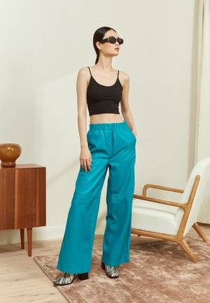 Calças largas turquesa com um acabamento texturizado, combinadas com um top curto preto justo e óculos de sol pretos, situadas num espaço interior minimalista.