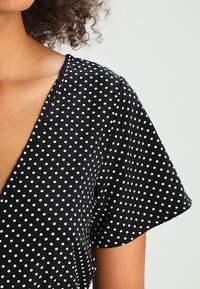 Mustapohjainen kangas, jossa on valkoisia pilkkuja, V-pääntie ja lyhyet, väljä hihat. Pehmeä tekstuuri ja sujuva pudotus.