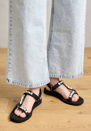Sandalias negras con diseño de tira en T, con gemas redondas y rectangulares, combinadas con jeans de pierna ancha de color azul claro sobre un suelo de madera.
