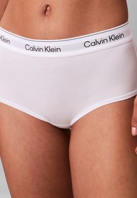 Od talii do górnej części ud osoby noszącej białe majtki Calvin Klein z elastycznym paskiem z logo na neutralnym tle.