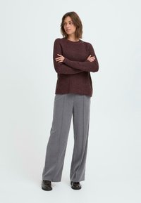 Maglione in cashmere color bordeaux a trama a coste, abbinato a pantaloni larghi grigi realizzati in un tessuto morbido. Calzature nere completano l'outfit.