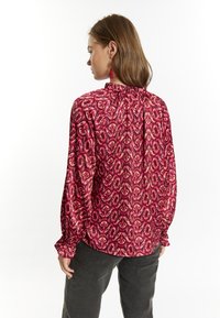 Blusa a maniche lunghe di un vivace rosa con un intricato motivo a foglie. Caratterizzata da un collo arricciato e polsini elastici. Tessuto liscio e leggero.