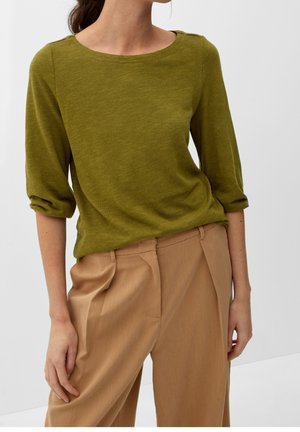 Kobieta ubrana w oliwkowo-zieloną bluzkę z długim rękawem z podwiniętymi mankietami oraz beżowe, dopasowane spodnie z wysokim stanem, stojąca na jednolitym tle.