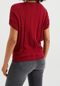 Osoba v červeném, krátkém ribbed topu a šedých džínách, stojící zády k fotoaparátu na jednorozměrném pozadí.