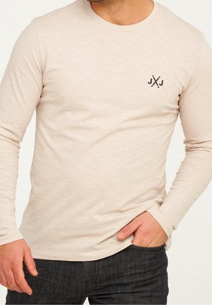 Chemise beige à manches longues avec petit logo noir "JXJ" sur la poitrine, portée avec un jean foncé, une main dans la poche, torse et bras visibles.