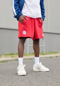 Shorts rouges avec poches, arborant un logo du Bayern Munich. Associés à des chaussettes blanches et des baskets Adidas crème avec des rayures grises. Veste bleue.