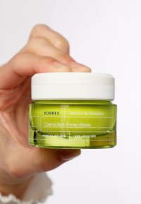 Grüner Glasbehälter der Korres Crème Anti-Pores, mit weißem Deckel und Textetikett, das den Säuregehalt und die Anwendung beschreibt, in einer Hand gehalten.