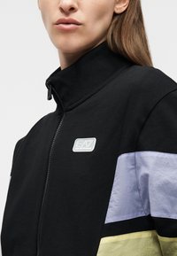 Femme portant une veste noire à fermeture éclair avec des panneaux de manches violet clair et jaune, et un petit logo EA7 Emporio Armani sur la poitrine.