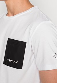 Bílé tričko s krátkým rukávem a černou kapsou, na které je napsáno "REPLAY" bílým písmem, kulatý výstřih a decentní detaily na ramenech.
