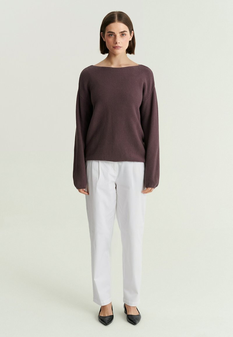 Femme aux cheveux courts et bruns portant un pull violet foncé à manches longues, un pantalon blanc et des ballerines noires pointues, debout devant un fond uni.