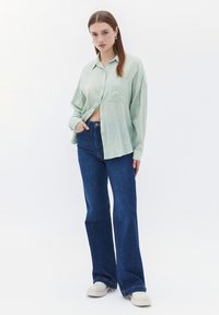 Lichtgroene button-up shirt met een zak, gecombineerd met donkerblauwe flared jeans. Model draagt witte schoenen en staat tegen een witte achtergrond.