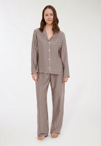 Donna in piedi a piedi nudi che indossa una camicia a maniche lunghe abbottonata abbinata e pantaloni larghi con strisce verticali nere e beige.