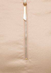 Beige tyg med en synlig metallisk dragkedja, som har en dragflik i guldton och sömdetaljer längs dragkedjans kant.