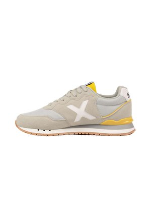 Zapatilla deportiva gris claro y beige con logo X blanco, detalle amarillo en el talón, diseño con cordones y suela de goma, mostrada de lado sobre fondo blanco.