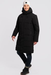 Abrigo de plumas negro con capucha, con cremallera y bolsillos con parche, de forma estilizada. Combinado con pantalones oscuros y botas de senderismo negras.