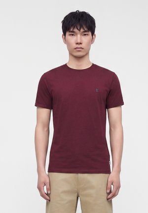 Ung mand med kort sort hår iført en ensfarvet vinrød kortærmet t-shirt og beige bukser, stående mod en lys baggrund.