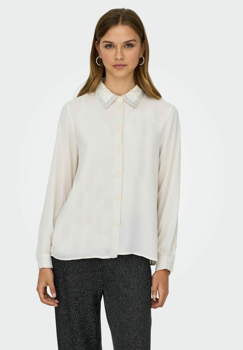 Chemise boutonnée crème clair avec un col et des manches longues. Le tissu semble lisse avec un léger éclat.