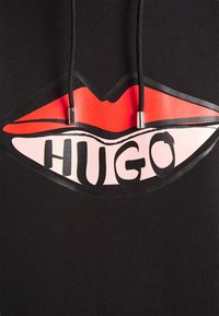 Černá mikina s kapucí, která má velký grafický design s červenými rty a slovem "HUGO" v tučných černých písmenech na růžovém pozadí. Texturovaná tkanina.