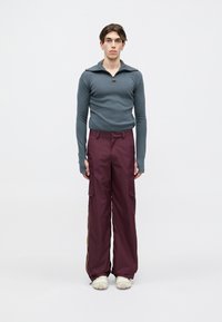 Šedé tričko s dlouhým rukávem a zipem u krku, rovně střižené, spojené s volnými vínově červenými kalhotami s bočními kapsami a žlutými pruhy po stranách.